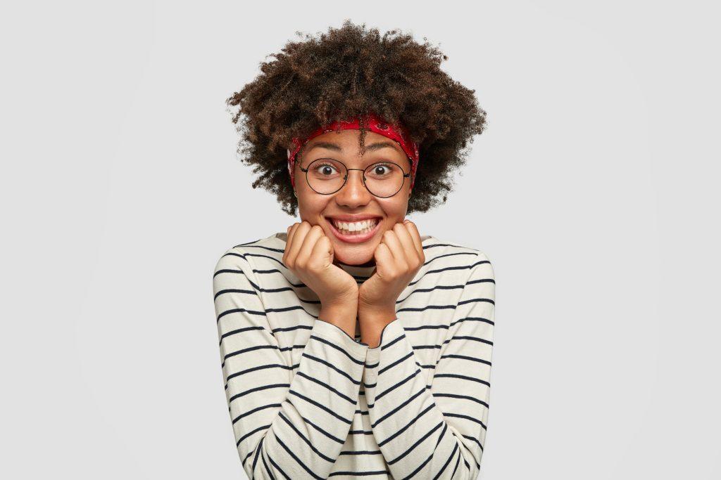 Delighted pleased African girl in transparent glasses has charming friendly smile, keeps both hands under chin, looks with amazement, hears positive story, models alone against white background.