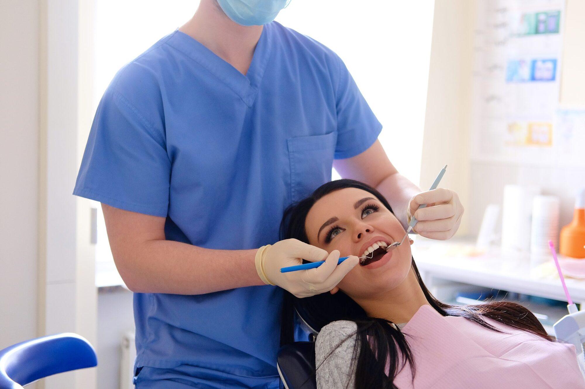 Dentist in blue uniform and white gloves cares on a sexy brunette woman, stand behind her.