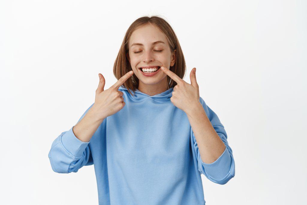 Portrait of happy women showing her white teeth after dentist whitening, pointing fingers at perfect smile, standing in blue t-shirt against white background